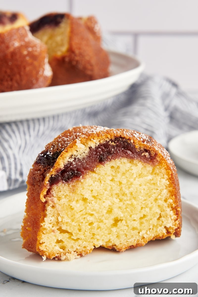 A perfectly sliced piece of Black Cherry Sour Cream Coffee Cake on a white plate, with the rest of the elegant Bundt cake visible in the background.