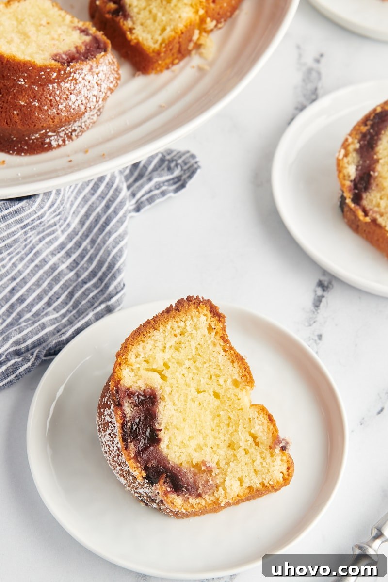 A close-up, appetizing shot of a slice of Black Cherry Sour Cream Coffee Cake on a white plate, with the remainder of the cake in soft focus behind it.