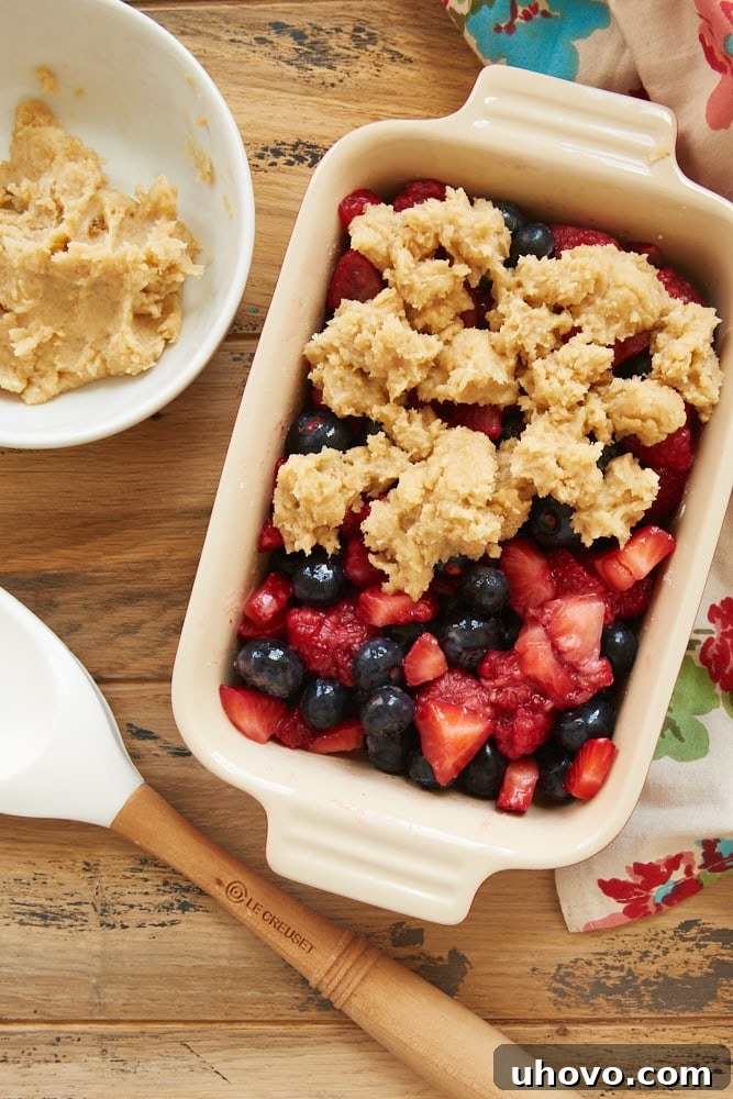 A close-up view of the Mixed Berry Cream Cheese Crisp during assembly, showcasing the vibrant berry mixture topped with dollops of the creamy, unbaked crisp topping, ready for the oven.