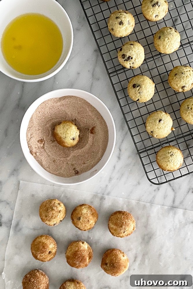 Hands carefully dipping a warm Mini Chocolate Chip Doughnut Muffin first into a bowl of melted butter, then gently rolling it in a bowl of cinnamon-sugar, creating the signature sweet coating.