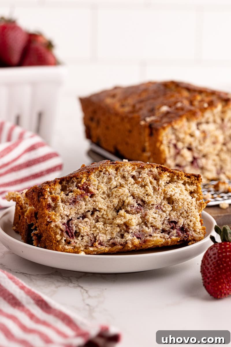 A beautifully baked slice of Strawberry Cream Cheese Bread on a white plate, with more bread and fresh strawberries gently blurred in the background, inviting you to take a bite.