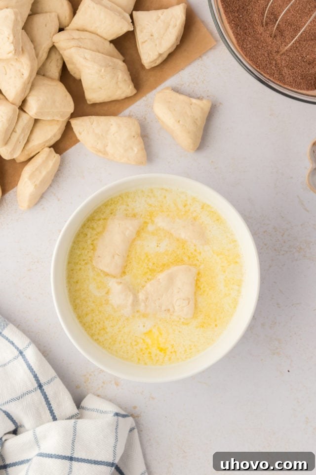 Overhead view of biscuit dough pieces being dipped into a bowl of melted butter, ensuring they are fully coated before moving to the sugar mixture.