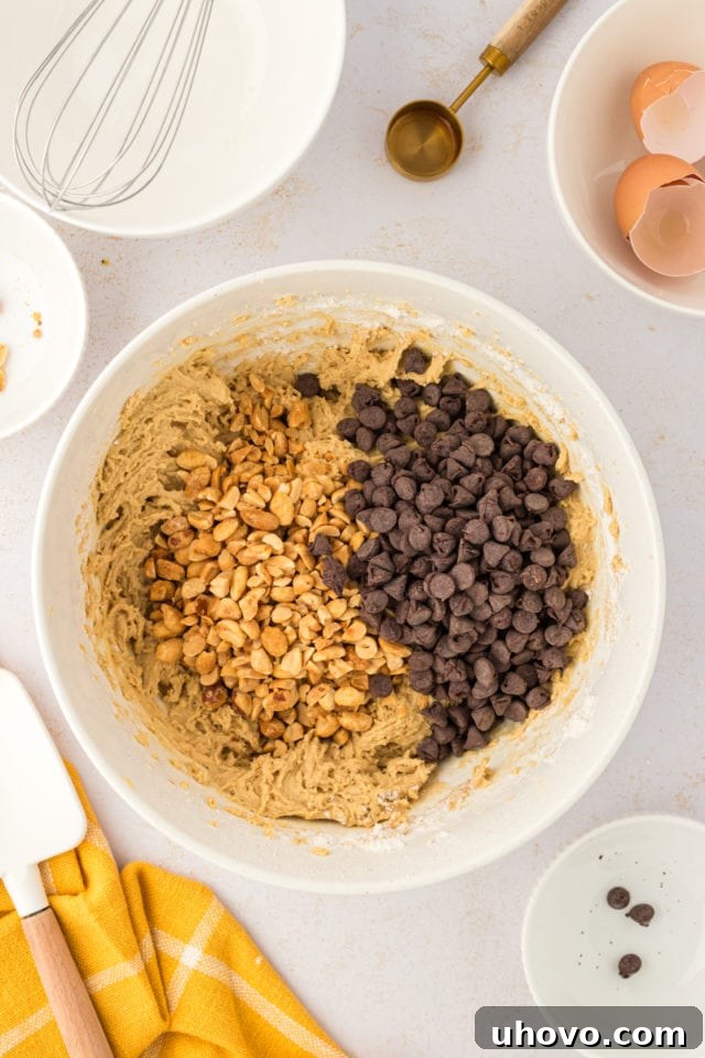An overhead view of crunchy peanuts and delicious chocolate chips being folded into the smooth toffee squares dough in a white mixing bowl.