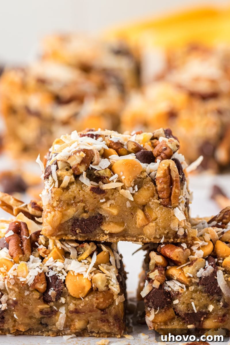 A stack of three golden brown toffee squares, showing their layered texture and generous toppings, with more delectable bars blurred in the background.