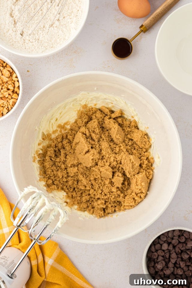 An overhead view of rich brown sugar being added to light, creamed butter in a pristine white mixing bowl, ready for blending.