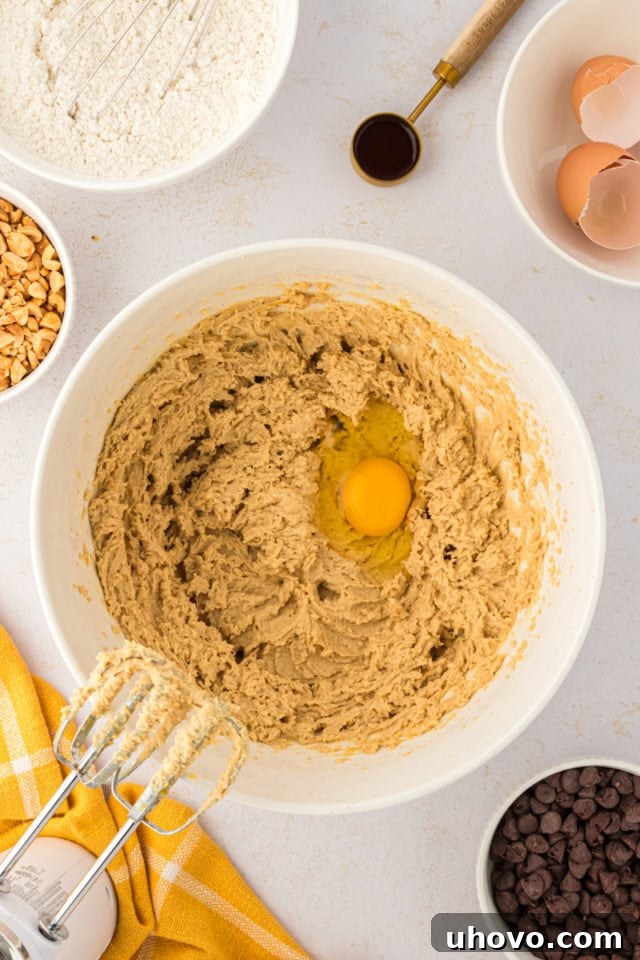 An overhead shot showing a single egg carefully added to the creamy mixture of butter and brown sugar in a white mixing bowl, ready to be incorporated.
