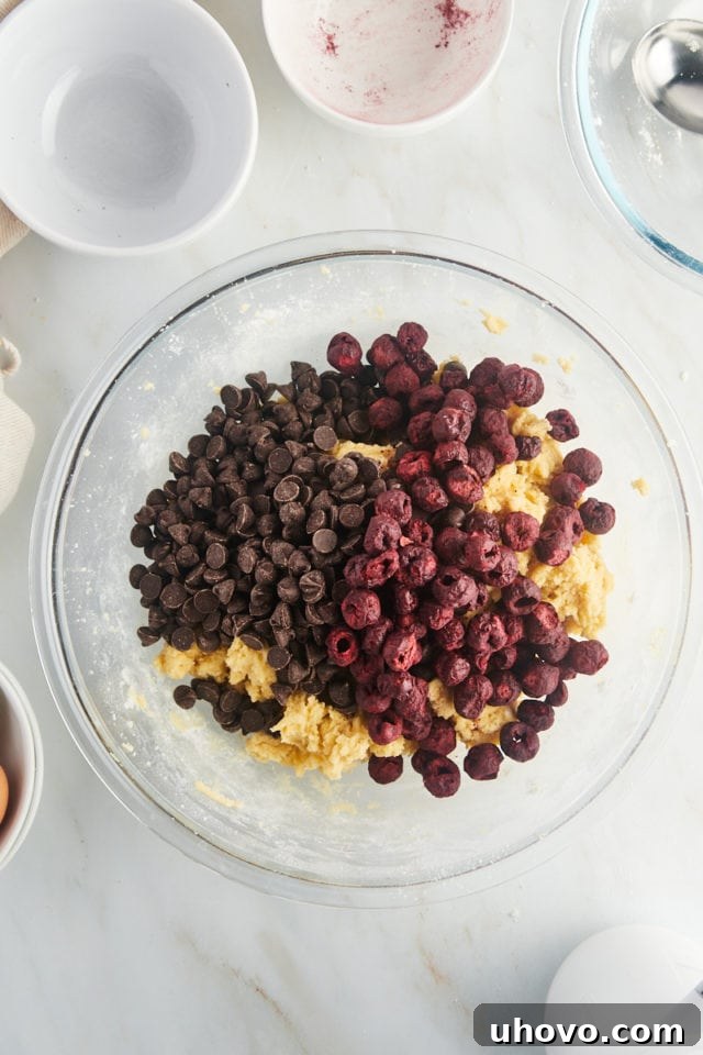 Semisweet chocolate chips and freeze-dried cherries layered on top of the cookie dough in a mixing bowl.