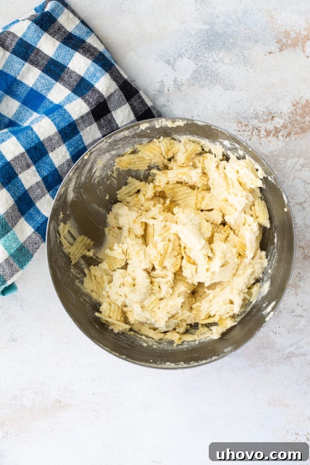 Overhead view of crushed potato chips being added to cookie dough in a metal mixing bowl, showcasing the unique ingredient combination.