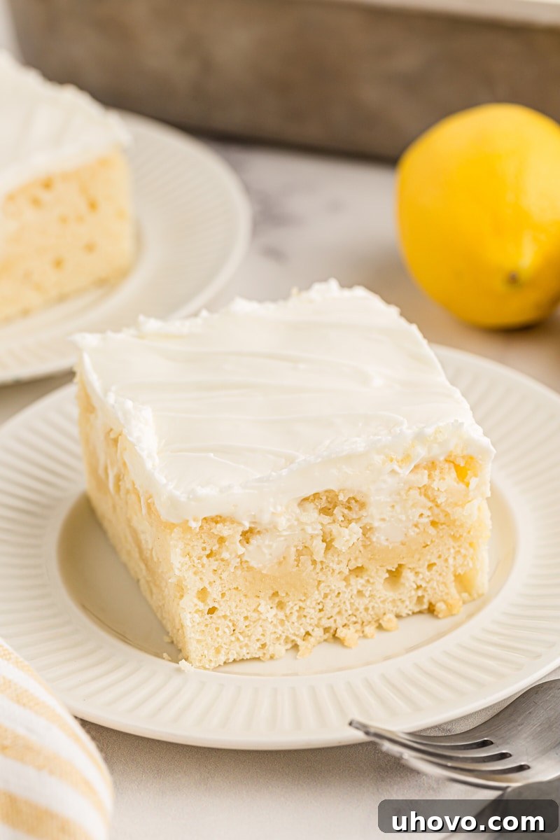 A pristine piece of lemon poke cake, artfully arranged on a white plate, invitingly showcasing its moist texture and perfect frosting.