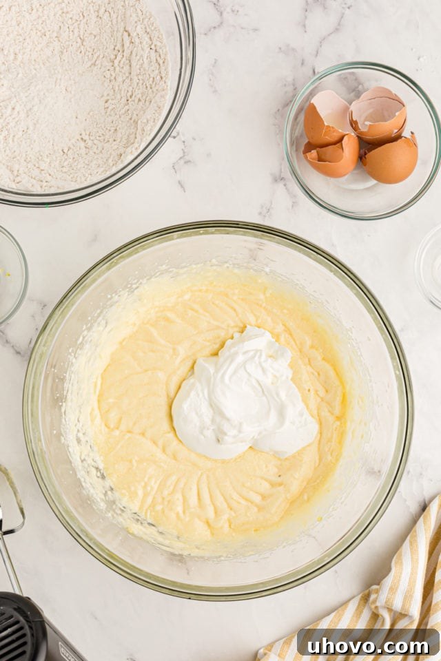 A vibrant overhead shot of sour cream being added to the cake batter, promising an exceptionally moist and tender texture for the finished lemon cake.