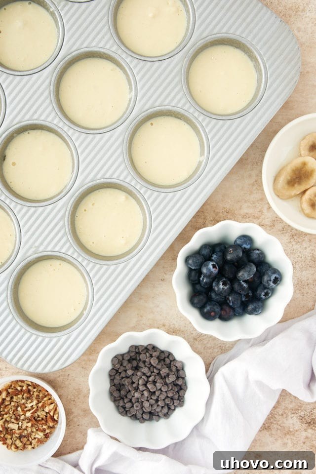 Pancake batter neatly poured into a standard muffin pan, with small bowls of various colorful toppings like fresh berries and chocolate chips arranged around it, ready for customization before baking.