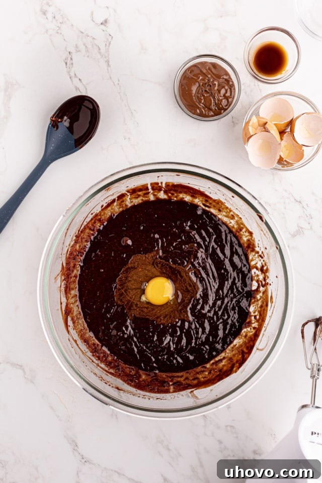 Overhead view of a large egg being added to the melted chocolate and sugar mixture in a mixing bowl.