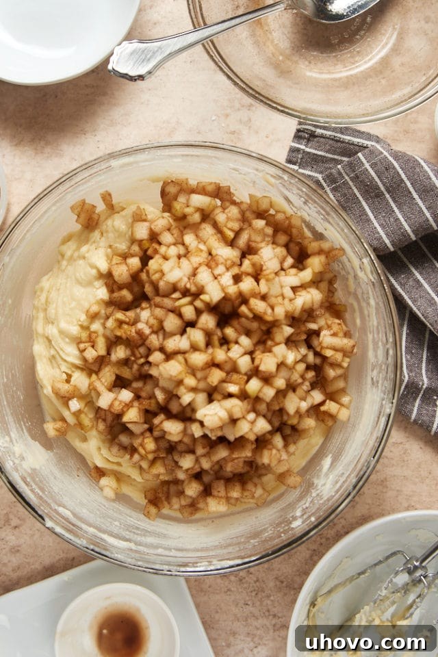 Chopped pears coated in cinnamon-sugar are gently folded into the cake batter in a glass mixing bowl, ready for baking.