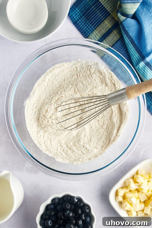 Overhead view of a whisk stirring dry ingredients (flour, sugar, baking powder, salt) in a glass mixing bowl.