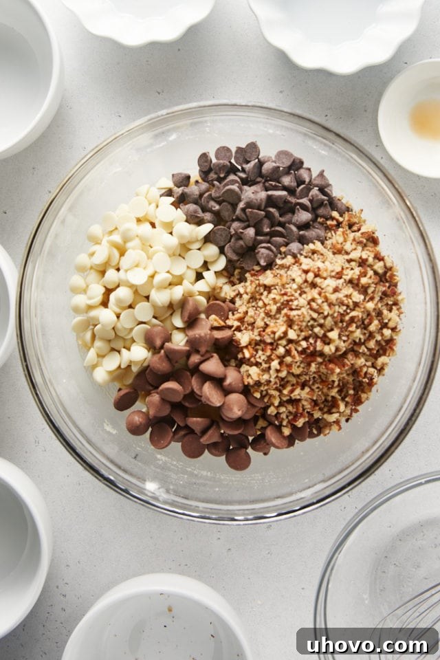 Overhead view of assorted chocolate chips and chopped nuts being added to the mixed cookie dough in a glass bowl.
