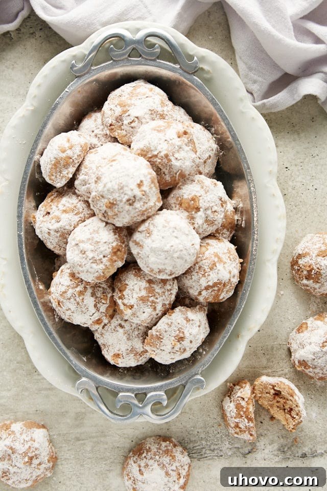An overhead view of numerous Nutty Spiced Snowball Cookies artfully arranged in an oval pewter tray, surrounded by more cookies on the surface, creating a bountiful and inviting presentation.