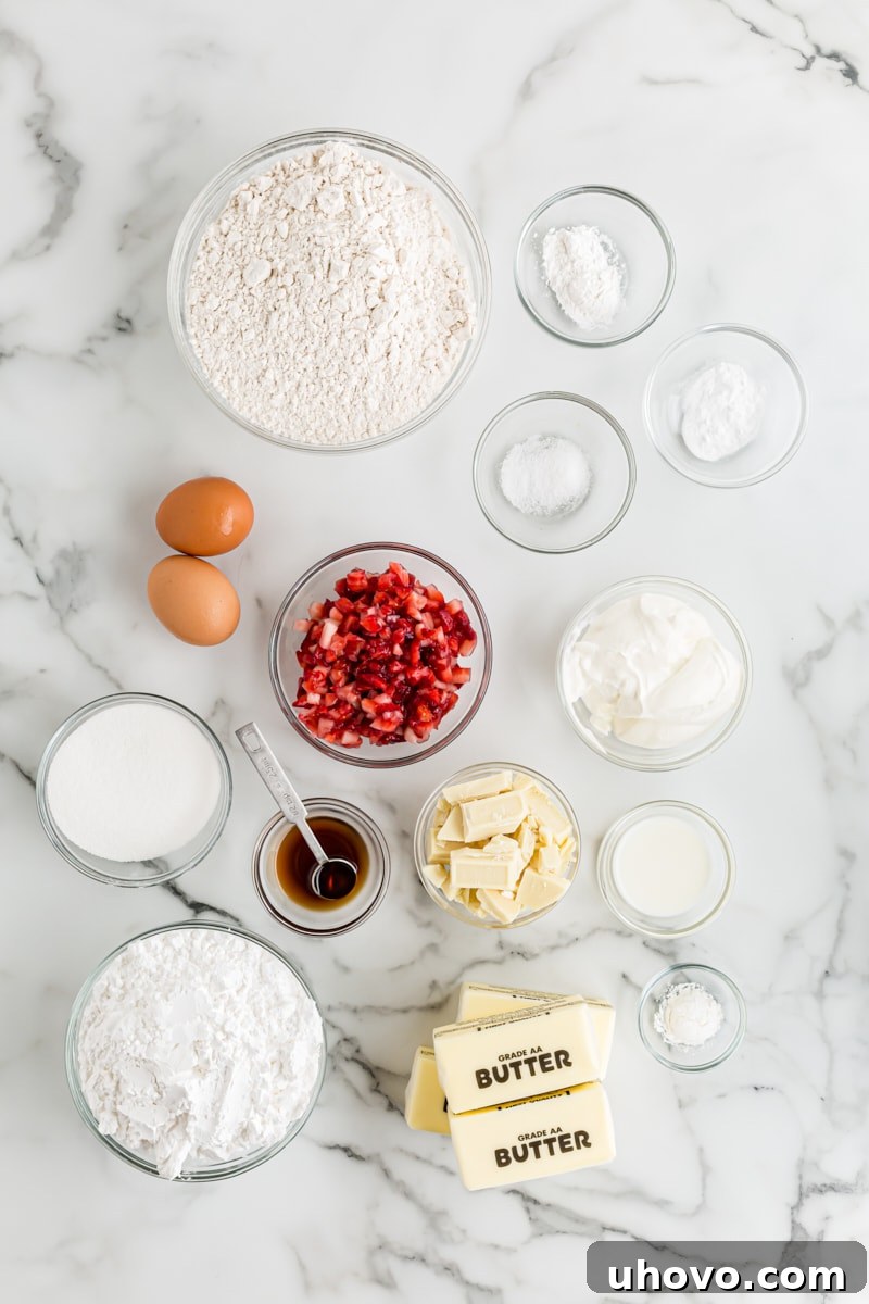 Overhead view of fresh ingredients for making strawberry cupcakes with white chocolate frosting, including flour, sugar, butter, eggs, sour cream, and a bowl of vibrant strawberries