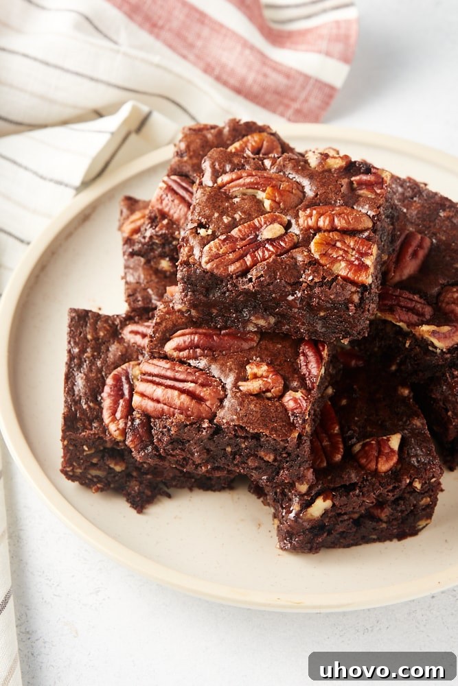 A generous pile of freshly baked Pecan Lovers' Brownies stacked on a beige ceramic plate, showcasing their rich, dark chocolate color and the visible pecan pieces on top.