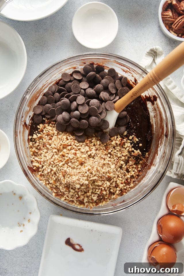 An overhead view of chopped pecans and chocolate chips scattered over the rich brownie batter, awaiting incorporation.