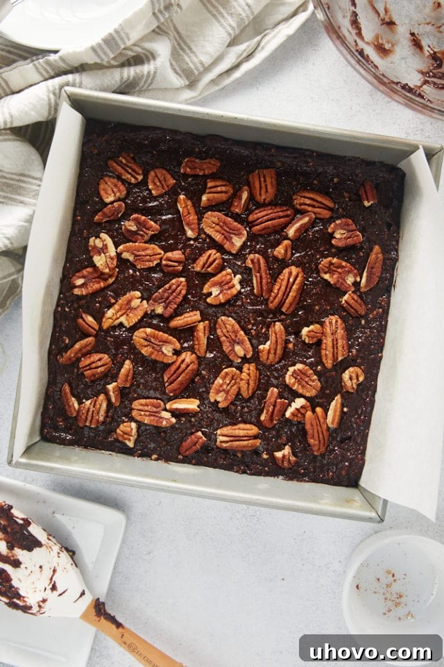 Overhead view of pecan halves and pieces generously sprinkled over the brownie batter in a parchment-lined baking pan, adding a decorative and nutty topping.