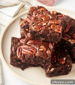 A generous pile of freshly baked Pecan Lovers' Brownies stacked on a beige ceramic plate, showcasing their rich, dark chocolate color and the visible pecan pieces on top.