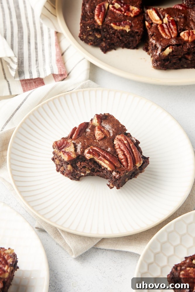 A single Pecan Lovers' Brownie with a bite taken out, resting on a white and beige plate, revealing its dense, fudgy interior and visible pecan pieces.