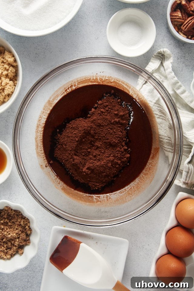 An overhead shot showing unsweetened cocoa powder being added to the melted chocolate and butter mixture in a glass bowl.