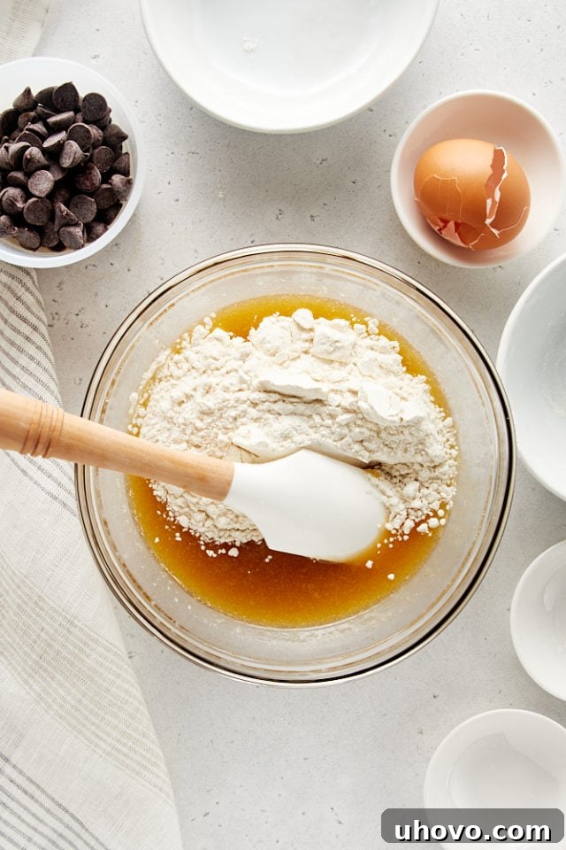 Overhead view of the dry flour mixture being added to the wet butter and sugar mixture in a glass bowl, beginning the final stage of cookie dough preparation.