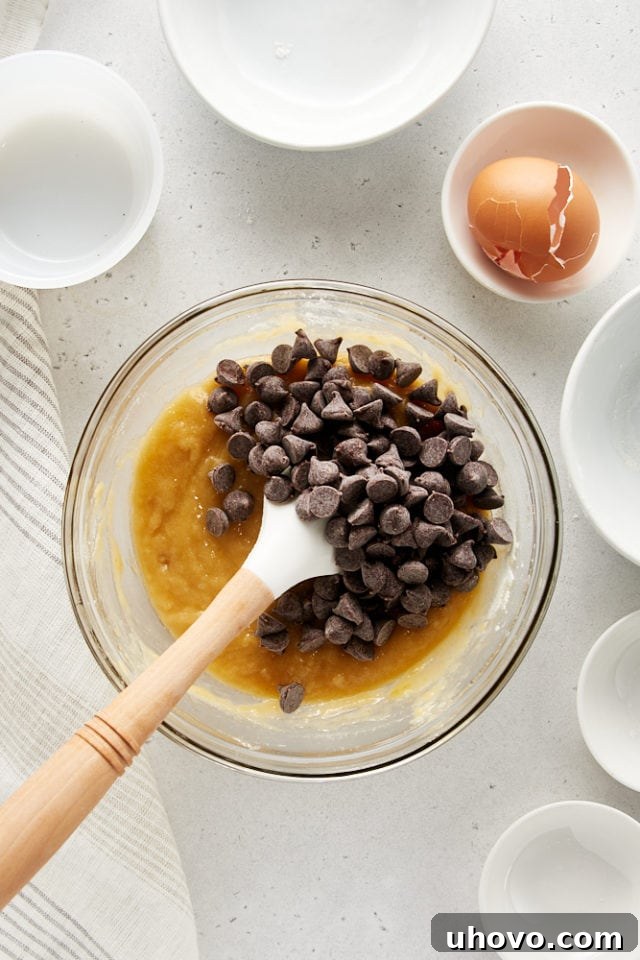 Overhead view of semisweet chocolate chips being folded into the prepared cookie dough in a glass bowl, ensuring even distribution for molten pockets of chocolate.