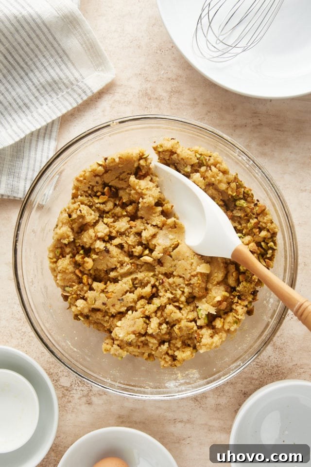 The cookie dough, now thoroughly mixed with vibrant green pistachios, sits in a glass mixing bowl, showcasing its readiness for chilling.