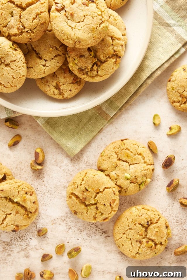 A beautiful arrangement of freshly baked Brown Butter Pistachio Cookies on a beige surface, with a few stacked on a beige plate, highlighting their perfect golden hue and pistachio flecks.
