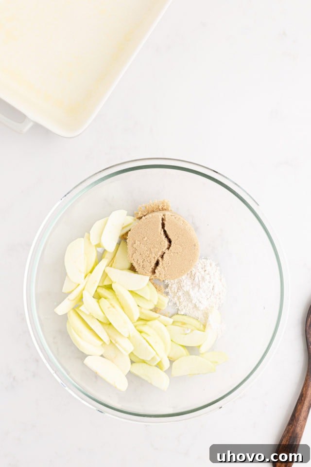 Overhead view of sliced apples, flour, and brown sugar being mixed in a large glass bowl.