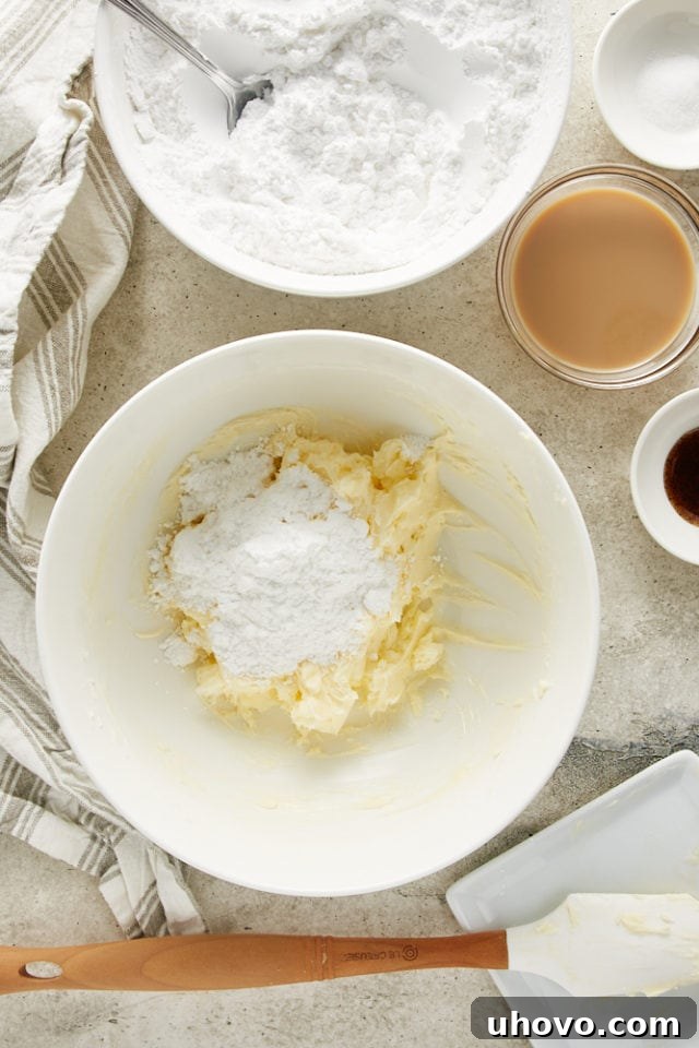 Confectioners' sugar being gradually added to creamed butter in a white mixing bowl, a key step in making smooth frosting.