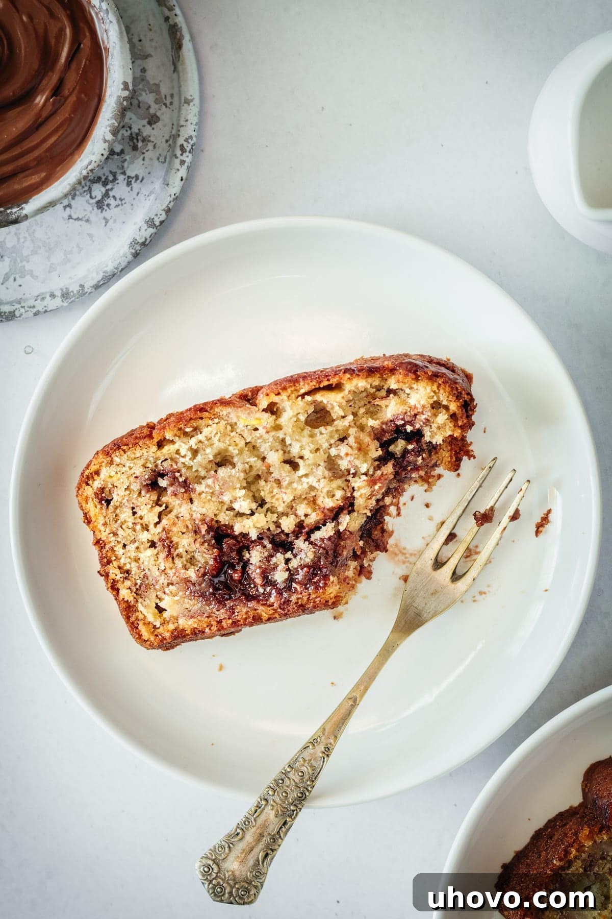 A perfectly sliced piece of Nutella Banana Bread on a white plate with a fork, showing beautiful chocolatey Nutella swirls within the incredibly moist banana bread, with a corner already enjoyed.