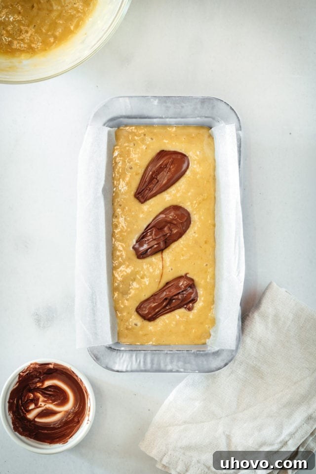 Overhead view of generous dollops of Nutella artfully added onto the first layer of banana bread batter in the loaf pan, poised for swirling to create beautiful ribbons.
