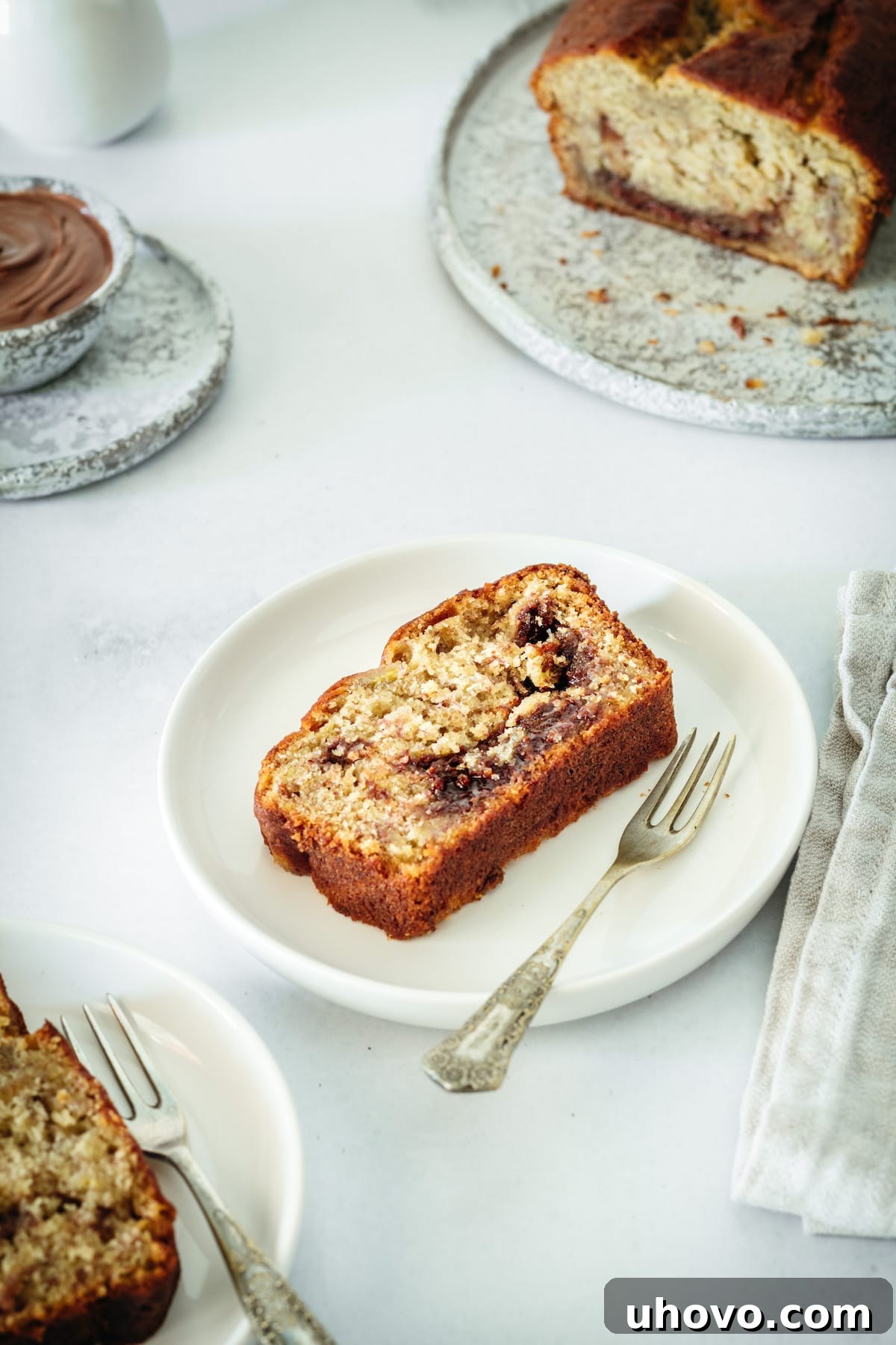 A delicious slice of Nutella Banana Bread on a white plate with a fork, with the remaining loaf beautifully blurred in the background, inviting you to enjoy its moist texture and rich Nutella swirls.