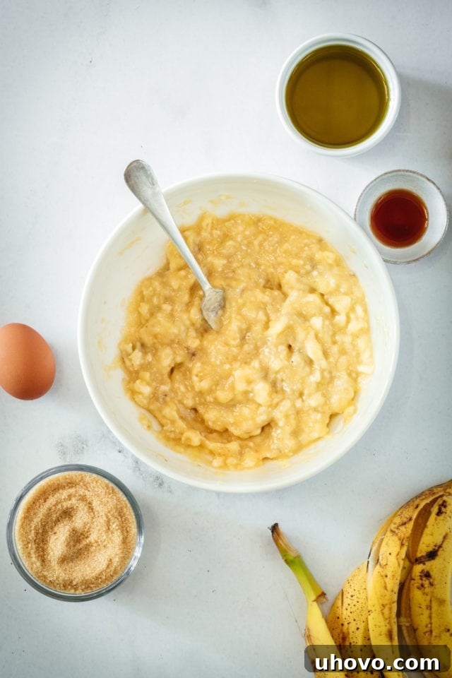 Overhead view of the mashed banana mixture in a mixing bowl with a spoon, showing the beginning stages of combining wet ingredients with the sugar.
