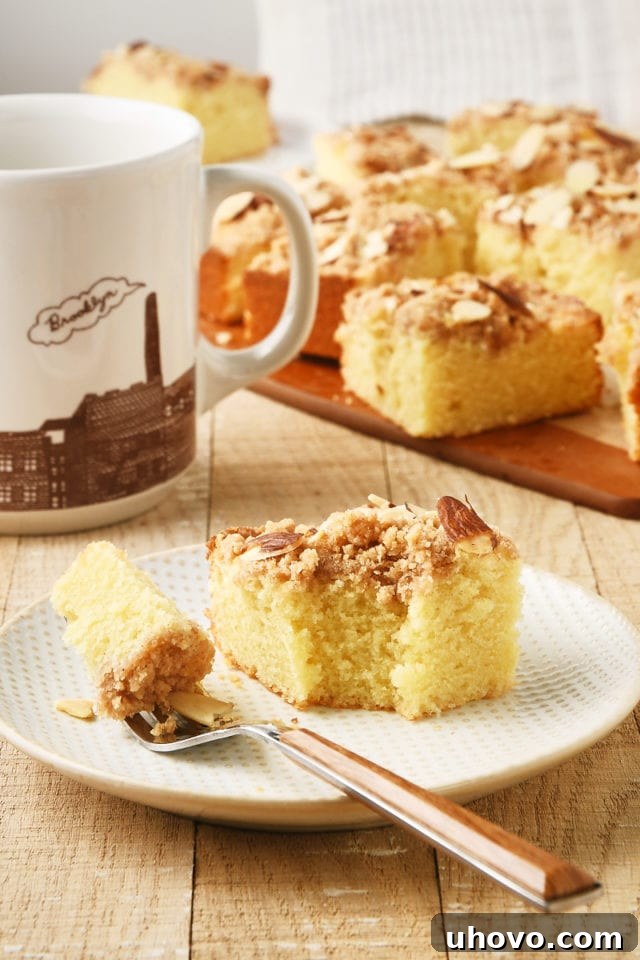 A close-up of a slice of Almond Crumb Cake on a white plate with a bite taken, a fork, and a coffee cup in the background.