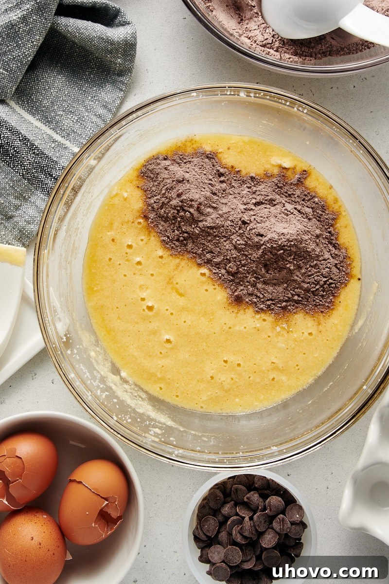 An overhead view capturing the moment dry ingredients are being added to the wet mixture in a glass bowl, a crucial step in preparing the Chocolate Loaf Cake batter.