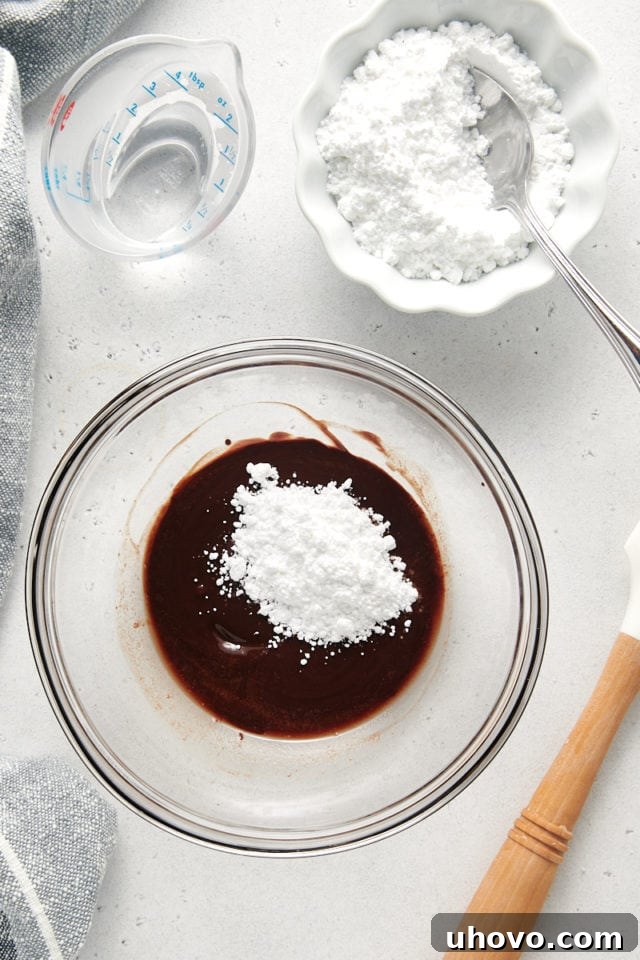 An overhead view of confectioners' sugar being gently incorporated into a bowl of melted butter and unsweetened chocolate, forming the rich chocolate glaze.