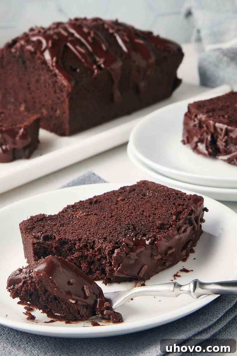 A single, delectable slice of Chocolate Loaf Cake with glaze, elegantly presented on a white plate, with the rest of the beautifully baked loaf blurred in the background.