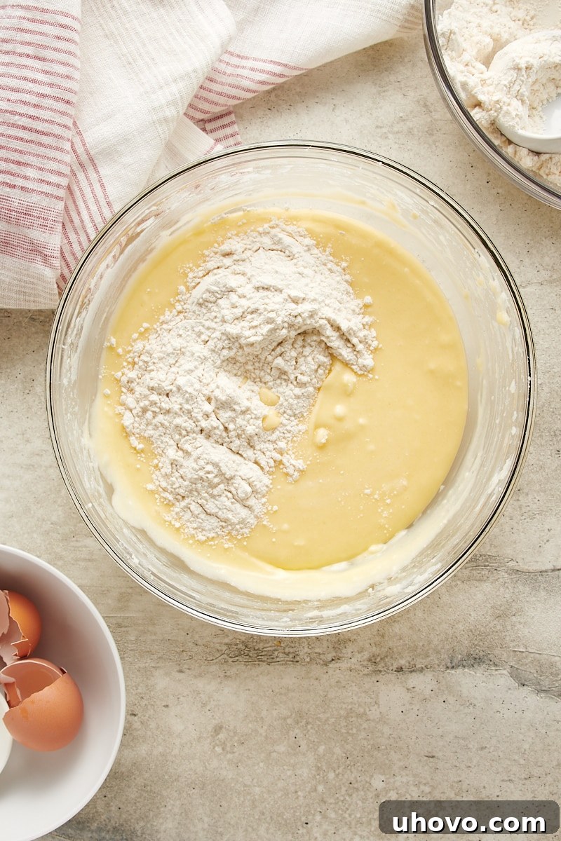 Overhead view showing the dry flour mixture gradually being added to the wet cream cheese dough in a glass bowl.