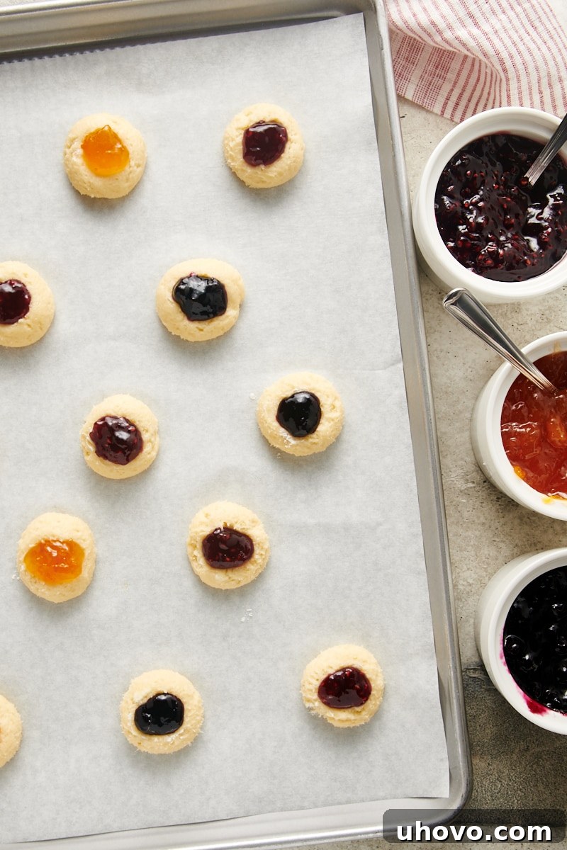 A baking sheet filled with Cream Cheese Thumbprint Cookies, each indentation generously filled with a variety of colorful jams, ready to be baked.