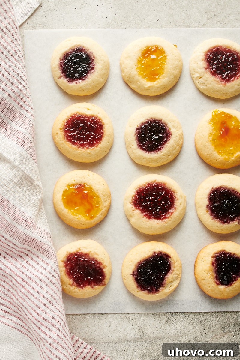 Overhead view of freshly baked Cream Cheese Thumbprint Cookies on white parchment paper, showcasing their golden edges and vibrant jam centers.