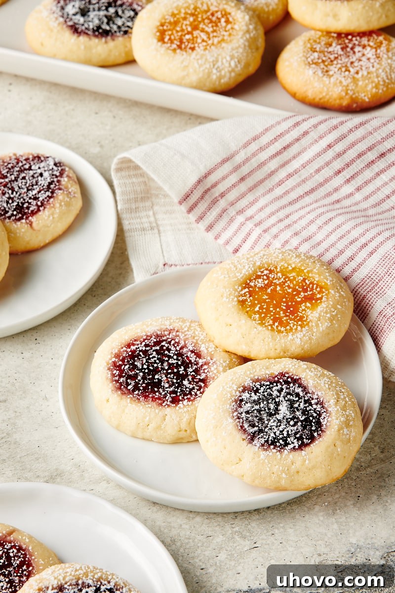 Three jam-filled Cream Cheese Thumbprint Cookies presented on a small white plate, with a soft-focused background of more cookies, highlighting their delicate centers.