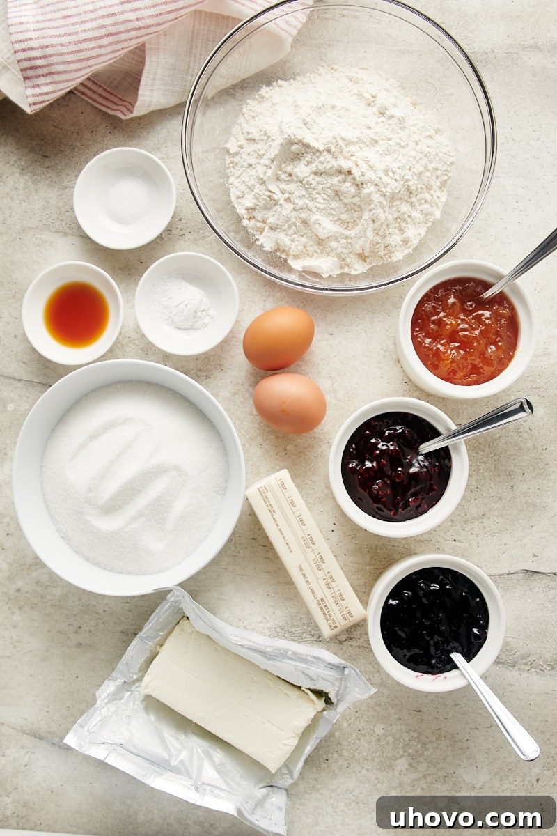 Overhead view of essential ingredients for Cream Cheese Thumbprint Cookies laid out on a kitchen counter: flour, butter, cream cheese, sugar, eggs, vanilla, and various jars of jam.
