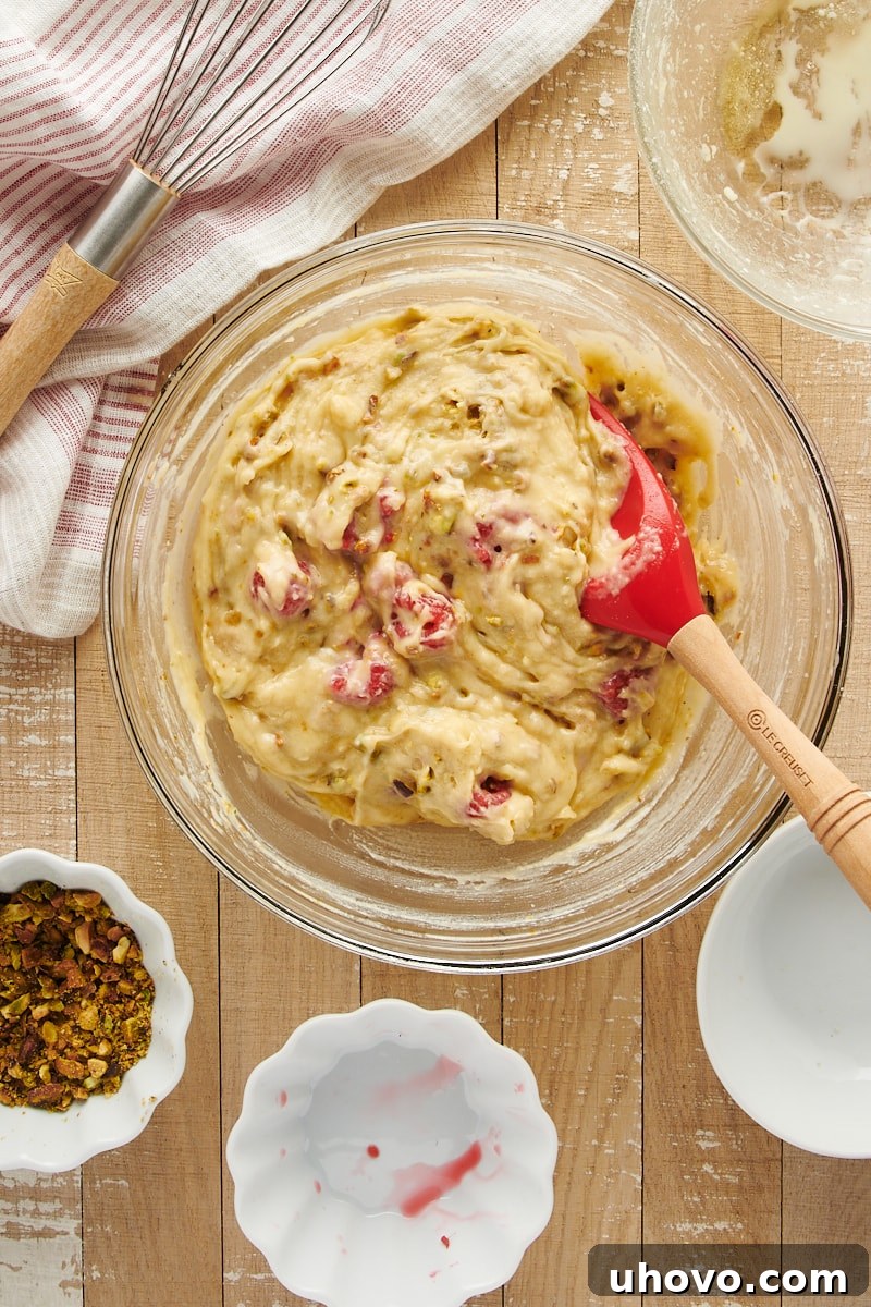 Overhead view of the fully mixed Raspberry Pistachio Muffin batter in a glass mixing bowl, with raspberries and pistachios evenly distributed throughout.