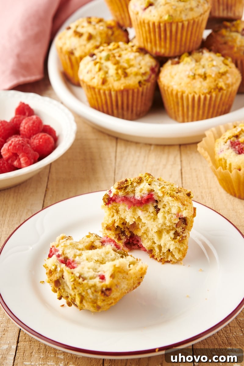 Two halves of a Raspberry Pistachio Muffin on a white plate with more whole muffins blurring in the background, showing the moist interior and colorful inclusions.