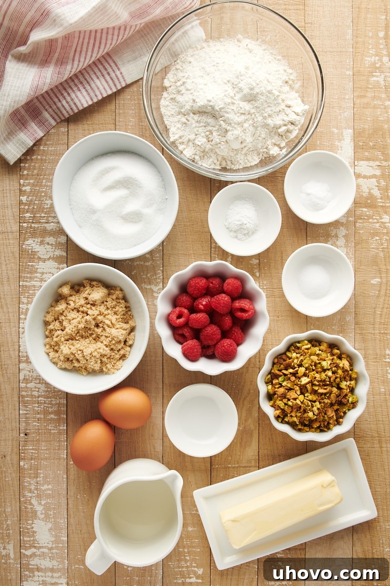 Overhead view of various fresh ingredients for Raspberry Pistachio Muffins laid out on a clean surface, including flour, sugars, butter, milk, eggs, raspberries, and pistachios.