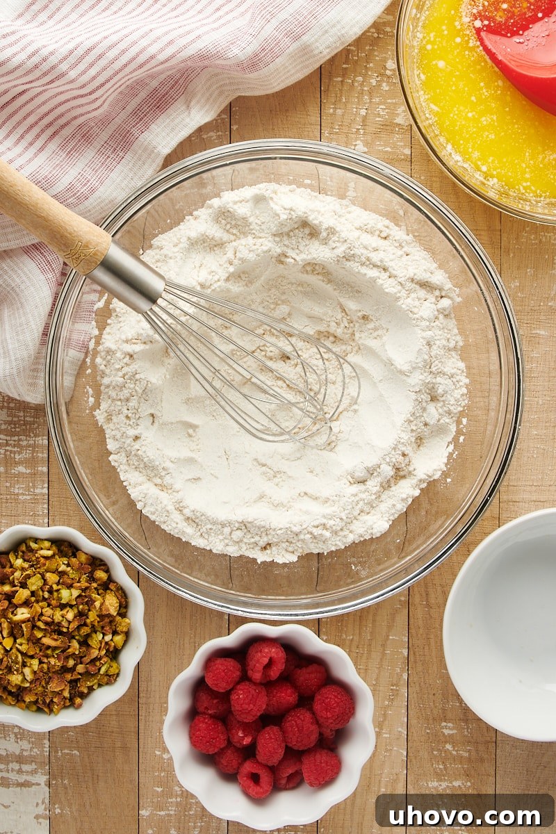 Overhead view of mixed dry ingredients for Raspberry Pistachio Muffins in a glass mixing bowl, showing the flour, sugars, and leavening agents blended.